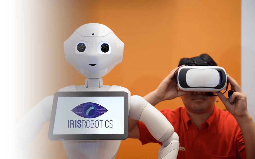 The Pepper robot with the IRIS Robotics logo on its tablet, next to a man wearing a VR headset