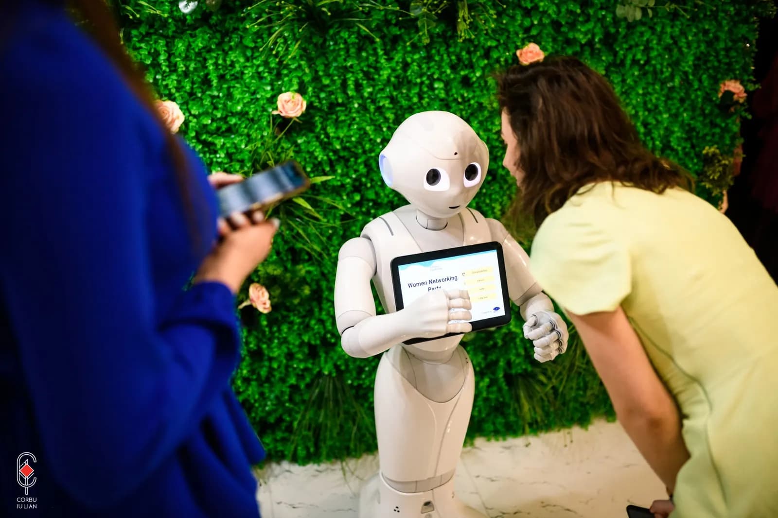 Pepper zeigt einer Besucherin das Menü auf seinem Tablet, vor einer grünen Wand mit Blumen